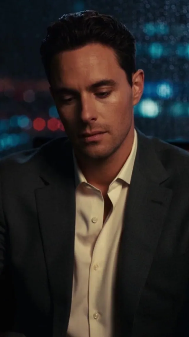 A fictional man in a suit, looking down in a rain-lit office